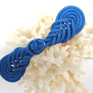 May include: A handcrafted blue rope decorative knot, featuring intricate weaving and a symmetrical design. The nautical-themed knot is displayed against a backdrop of off-white coral, creating a striking contrast. This unique piece is perfect for nautical decor.