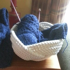 May include: A white rope basket filled with blue yarn and knitting needles. The basket is sitting on a wooden table.
