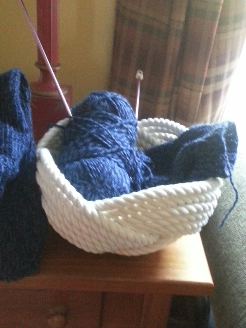 May include: A white rope basket filled with blue yarn and knitting needles. The basket is sitting on a wooden table.