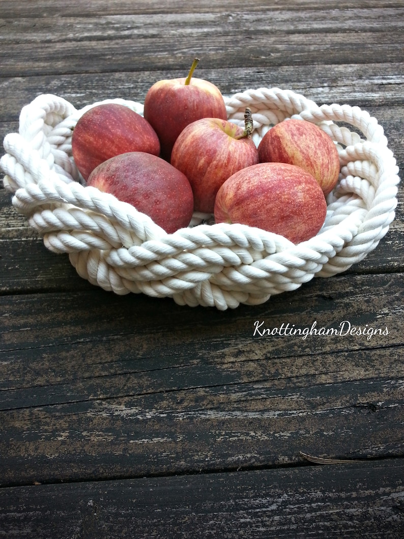 May include: A white rope bowl filled with red apples. The bowl is made of thick, braided rope and has a natural, rustic look. The apples are arranged in a circular pattern, with the stems facing outwards.