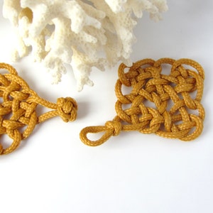 May include: Two mustard yellow macrame knot ornaments with a square and a teardrop shape. The ornaments have a loop at the top for hanging.