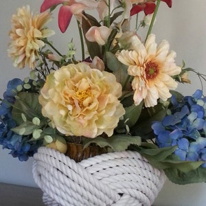 May include: A white rope basket filled with a variety of artificial flowers, including pink lilies, yellow roses, and blue hydrangeas.