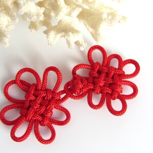 May include: Two red Chinese knot charms in the shape of flowers. The knots are tied with red string and are connected together.