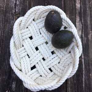 May include: White rope bowl containing two avocados. The bowl is crafted from thick white rope woven into a basket-like design. The avocados are dark green and have a smooth, glossy skin.