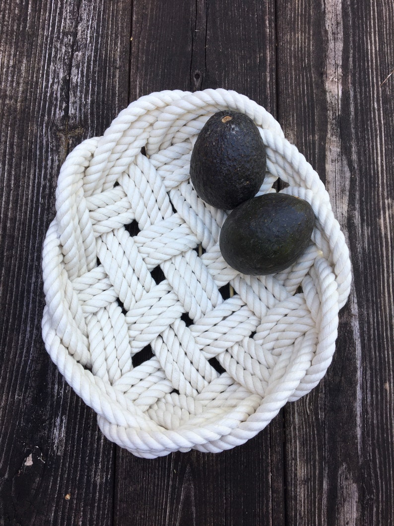 May include: White rope bowl containing two avocados. The bowl is crafted from thick white rope woven into a basket-like design. The avocados are dark green and have a smooth, glossy skin.