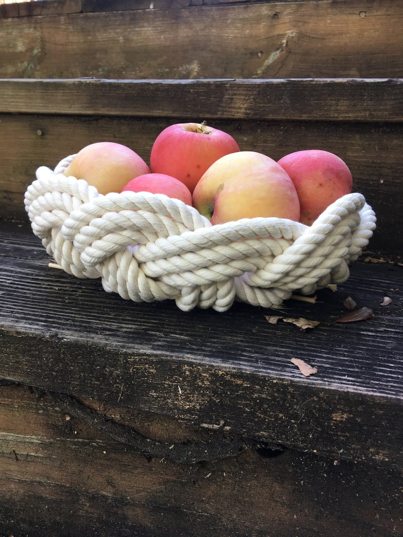 May include: A white rope bowl filled with red and yellow apples. The bowl is sitting on a wooden surface.