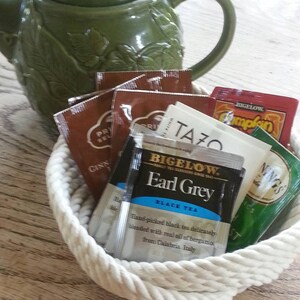 May include: A white rope basket filled with a variety of tea bags, including Bigelow Earl Grey black tea, Tazo tea, and a green tea bag with the word "NINGS" on it. The basket is sitting on a wooden table next to a green ceramic teapot.