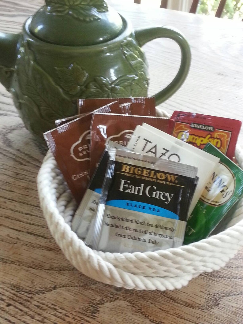 May include: A white rope basket filled with a variety of tea bags, including Bigelow Earl Grey black tea, Tazo tea, and a green tea bag with the word "NINGS" on it. The basket is sitting on a wooden table next to a green ceramic teapot.