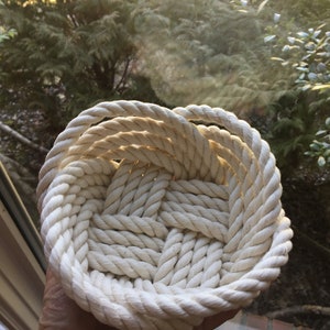 May include: A white rope basket woven into a circular shape. The basket is made of thick, natural rope and has a smooth, textured surface.