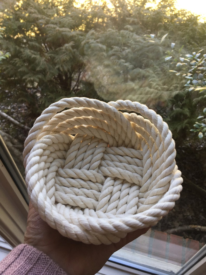 May include: A white rope basket woven into a circular shape. The basket is made of thick, natural rope and has a smooth, textured surface.