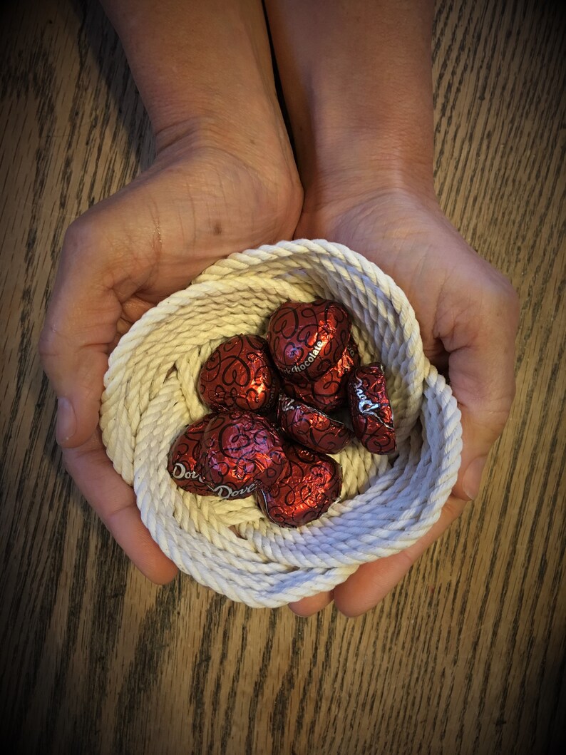 May include: A white rope bowl filled with red heart-shaped chocolates. The bowl is held in two hands.