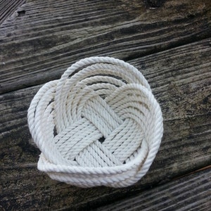May include: A white rope basket woven into a heart shape. The basket is sitting on a wooden surface.