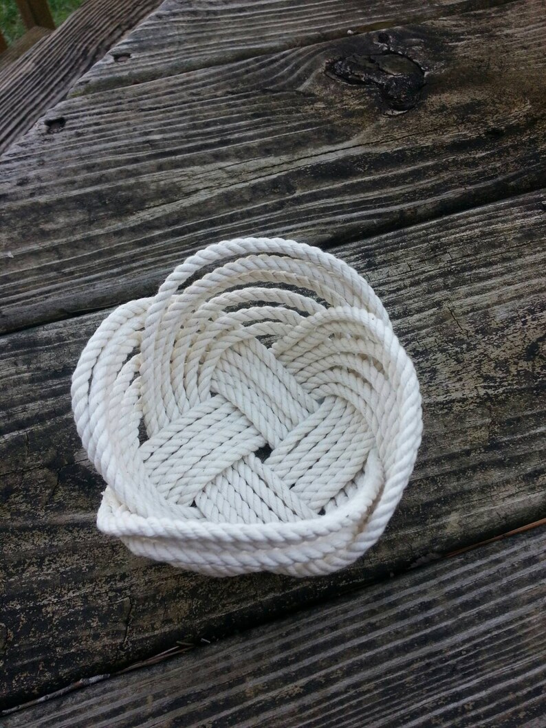 May include: A white rope basket woven into a heart shape. The basket is sitting on a wooden surface.