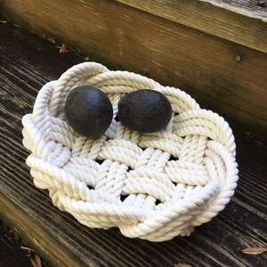 May include: A white rope bowl with two dark grey round objects inside. The bowl is made of thick rope and has a woven pattern. The bowl is sitting on a wooden surface.