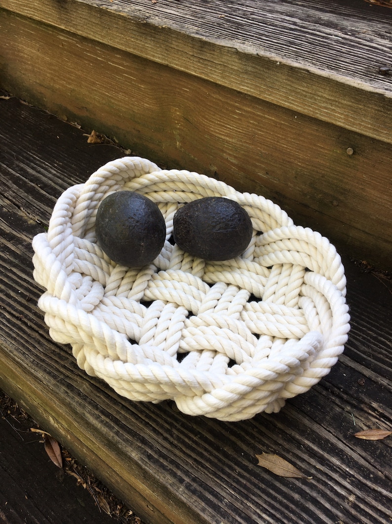 May include: A white rope bowl with two dark grey round objects inside. The bowl is made of thick rope and has a woven pattern. The bowl is sitting on a wooden surface.