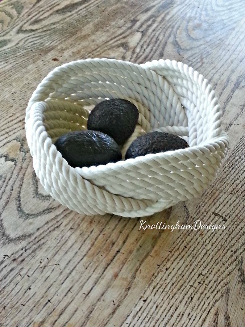 May include: A white rope bowl with three avocados inside. The bowl is made of thick, white rope and has a woven design. The avocados are dark green and have a smooth, shiny skin.