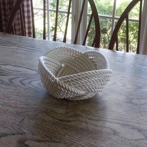 May include: A white rope bowl, woven in a knot pattern, sits on a wooden table.