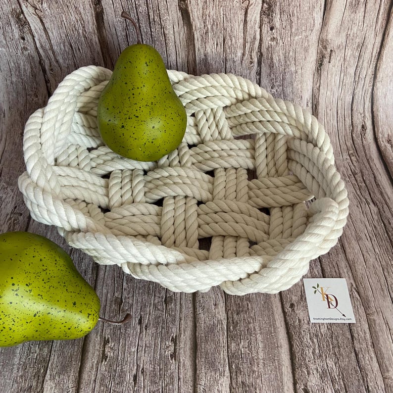 May include: A white rope woven basket with a pear inside. The basket is oval shaped and has a thick rope border. The basket is sitting on a wooden surface.