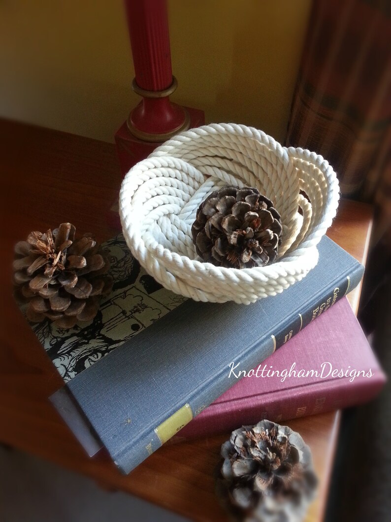 May include: A white rope bowl with a natural pine cone inside. The bowl is sitting on top of two books, one blue and one red. The text "KnottinghamDesigns" is visible on the red book.