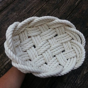 May include: A white rope basket woven in a lattice pattern. The basket is round and has a wide, flat base.