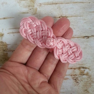 May include: A light pink, woven knot closure, likely for a garment or accessory. The closure is held in a hand, showcasing the intricate knotting pattern and the soft, pastel color. The background is a light wood grain.