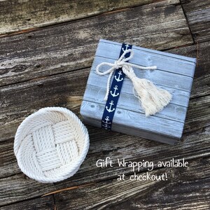 May include: A white rope basket with a woven pattern sits on a wooden surface next to a grey gift box with a blue ribbon and white tassels. The ribbon has a pattern of white anchors. The text "Gift Wrapping available at checkout!" is written on the wooden surface.
