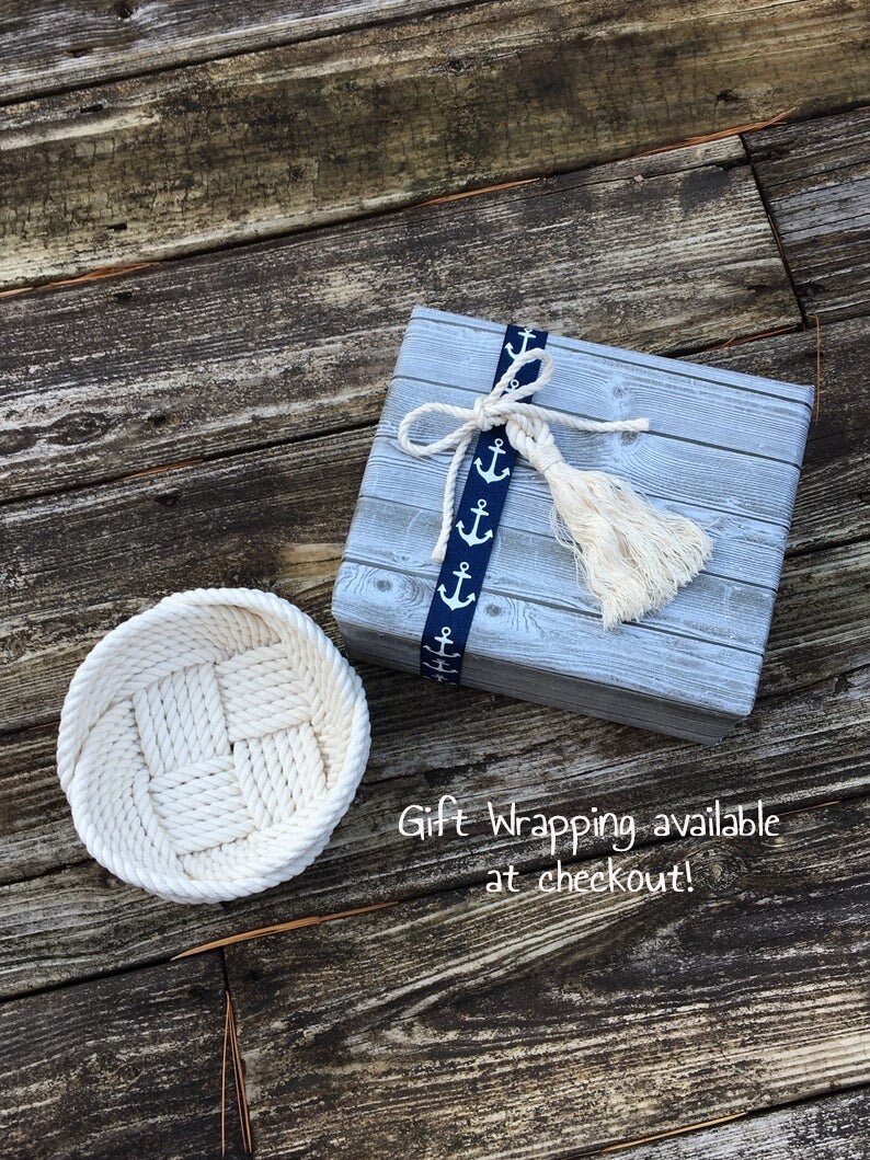 May include: A white rope basket with a woven pattern sits on a wooden surface next to a grey gift box with a blue ribbon and white tassels. The ribbon has a pattern of white anchors. The text "Gift Wrapping available at checkout!" is written on the wooden surface.