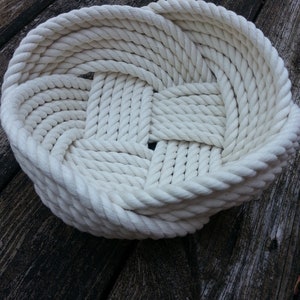 May include: A white rope basket woven in a circular shape. The basket is made of thick, white rope and has a natural, rustic look.