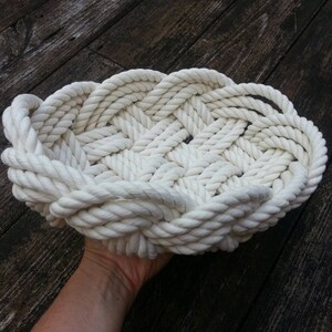 May include: A white rope basket woven into a circular shape. The basket is made of thick, white rope and has a slightly raised edge.