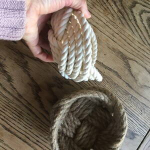 May include: Two rope baskets, one is white and the other is brown. The white rope basket is coiled up and the brown rope basket is open and ready to hold items.