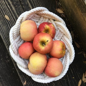 May include: A white rope basket filled with red and yellow apples. The basket is on a wooden surface.