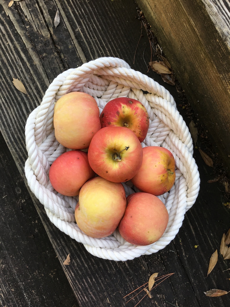 May include: A white rope basket filled with red and yellow apples. The basket is on a wooden surface.