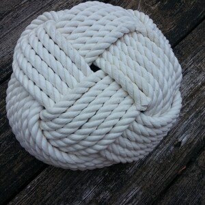 May include: A white rope tied into a large knot, resembling a ball. The rope is thick and has a coarse texture. The knot is on a wooden surface.