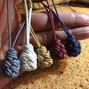 May include: Five small, knotted cord ornaments in different colors: gray, white, gold, burgundy, and navy blue. The cords are tied together and held in a hand.