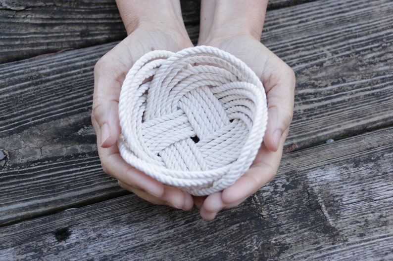 May include: A white rope bowl, woven in a circular pattern, is held in the hands of a person. The bowl is made of natural rope and has a simple, rustic design.
