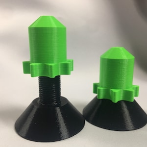 May include: Two green plastic 3D printed objects with a star-shaped base and a screw-like shaft. The larger object has a taller, more rounded top. The smaller object has a shorter, more conical top. Both objects are on a white surface.