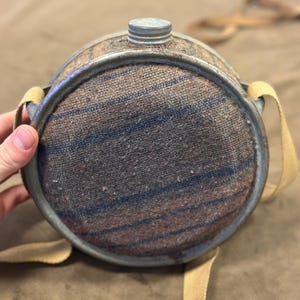 Peut inclure: Gourde vintage en métal avec un corps recouvert de tissu dans des tons bruns et bleus. La gourde a un bouchon en métal et une sangle en toile. La gourde est de forme circulaire.