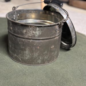 Puede incluir: Un cubo de metal vintage con tapa y asa. El cubo es verde y tiene un aspecto desgastado y rústico.