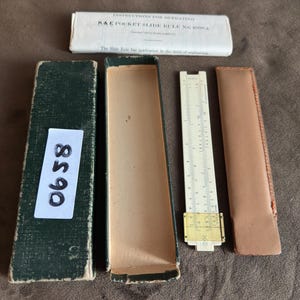 Pode incluir: Uma régua de cálculo vintage aberta com sua caixa e instruções. A régua de cálculo é esbranquiçada com marcações pretas e uma moldura de latão. A caixa é verde e as instruções estão em uma manga de papel branco.