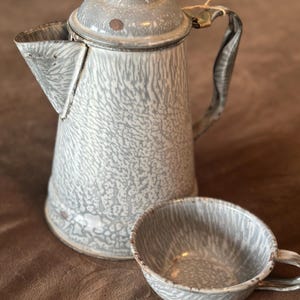 May include: A vintage gray and white speckled enamelware coffee pot with a lid and a matching cup. The coffee pot has a black handle and a spout. The cup has a handle.