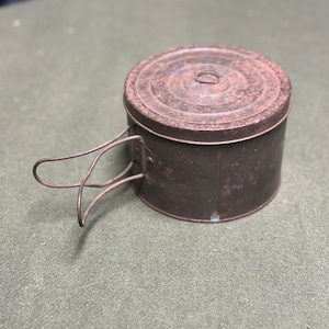 Peut inclure: Un récipient en métal vintage rouillé avec un couvercle à charnière et une poignée en fil de fer. Le récipient est rond et a un couvercle légèrement bombé.