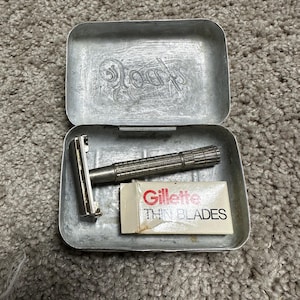 Peut inclure: Un rasoir argenté avec un paquet de lames Gillette Thin Blades dans un étui en métal argenté. L'étui porte le mot "Gillette" en relief sur le dessus.