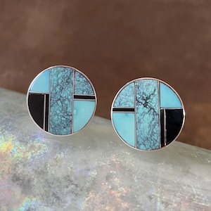 May include: A pair of round stud earrings with a silver border. Each earring features a mosaic design with turquoise, black, and light blue segments. The turquoise sections have a textured appearance, creating a striking contrast.