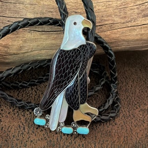 May include: A detailed bolo tie featuring an eagle design. The eagle has a white head, black and brown wings, and is accented with turquoise stones. The bolo tie has a black braided leather cord.