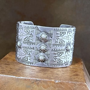 May include: A wide, silver cuff bracelet with intricate geometric and stylized bird designs. The cuff features a series of stamped patterns, including repeating bird motifs, triangles, and circular elements. The bracelet has a vintage or handcrafted aesthetic.