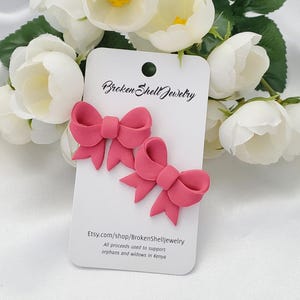 May include: Two pink polymer clay bow earrings on a white card with the text "Broken Shell Jewelry" and the website address "Etsy.com/shop/BrokenShellJewelry". All proceeds are used to support orphans and widows in Kenya.