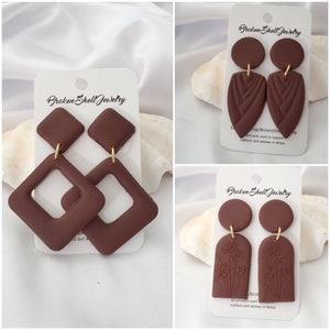 May include: Three pairs of brown clay earrings on white cards. The earrings are all different shapes and designs. The cards have the text "Broken Shell Jewelry" on them.