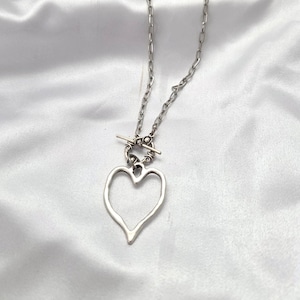 May include: Silver-toned necklace featuring a large, open heart pendant. The heart is suspended from a chain with a toggle clasp closure. The necklace is displayed on a white, textured background.