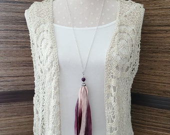 May include: A long silver chain necklace with a purple and pink tassel pendant. The tassel is made of silk fabric and has a gradient of color from purple to pink. The necklace is displayed on a white crocheted vest.