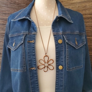 May include: A copper daisy necklace with a large, openwork pendant. The necklace is worn over a white shirt and a denim jacket.